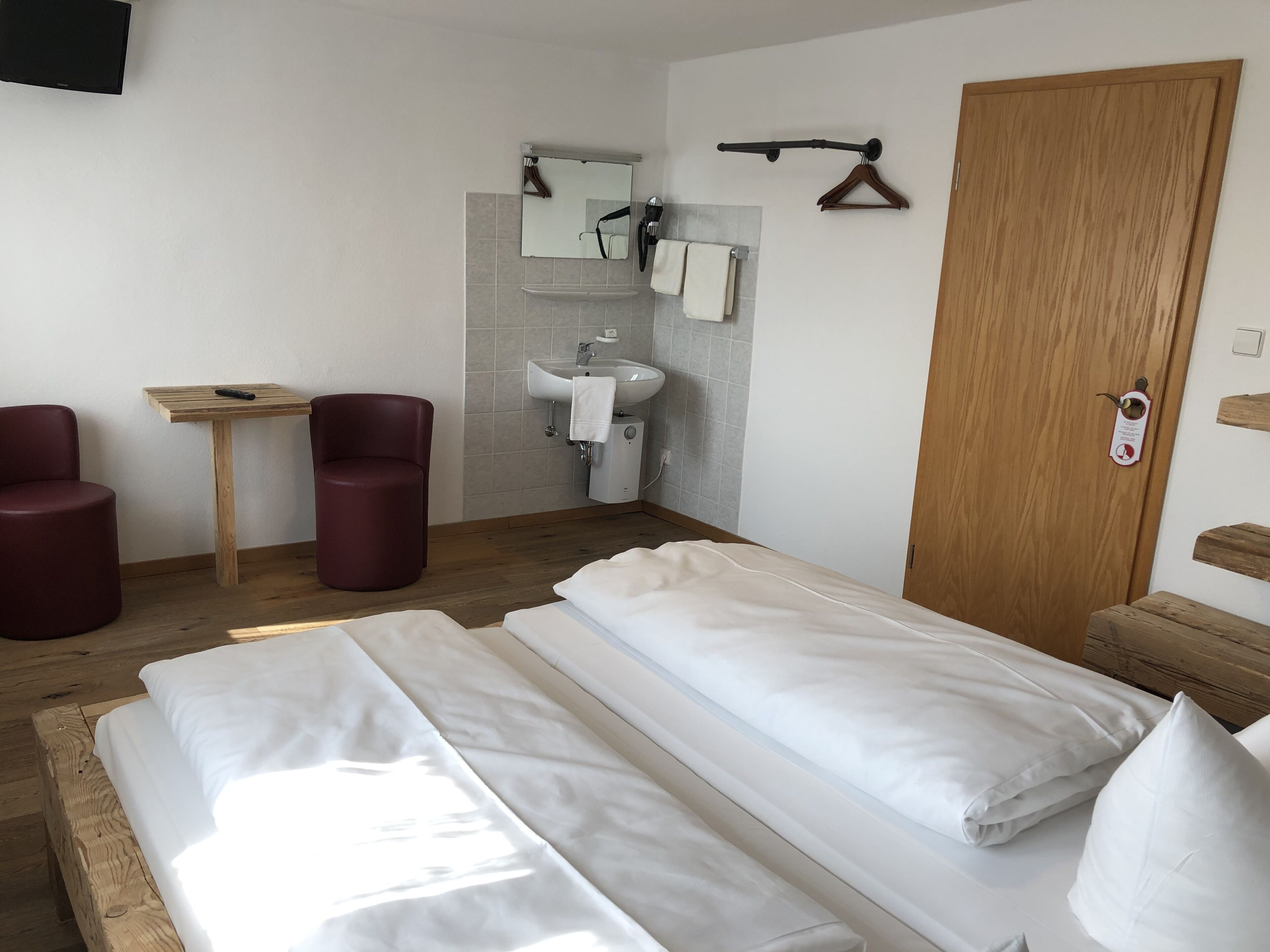 Une chambre d’hôtel avec deux lits, une table en bois, deux chaises rouges, une armoire en bois, un miroir, un porte-serviettes et un lavabo.