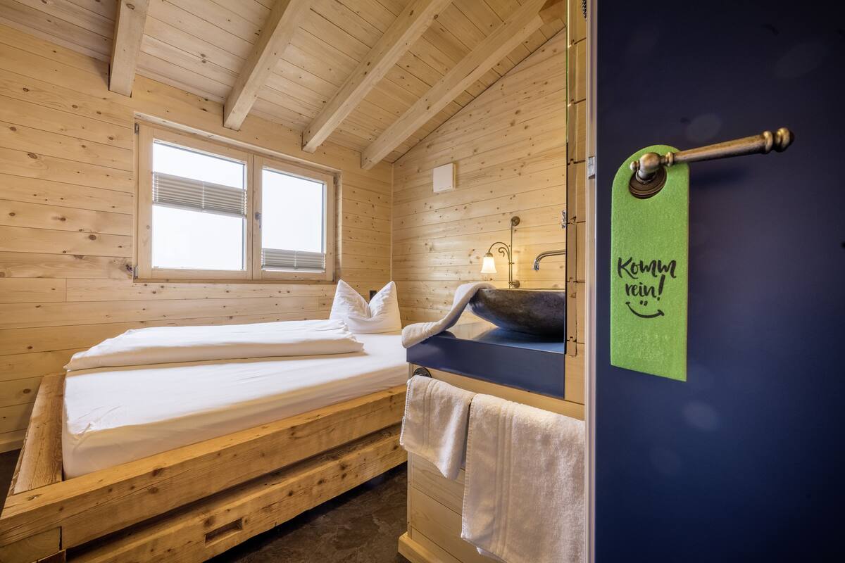 Een houten kamer met een bed, een raam, een wastafel en een handdoekhouder.