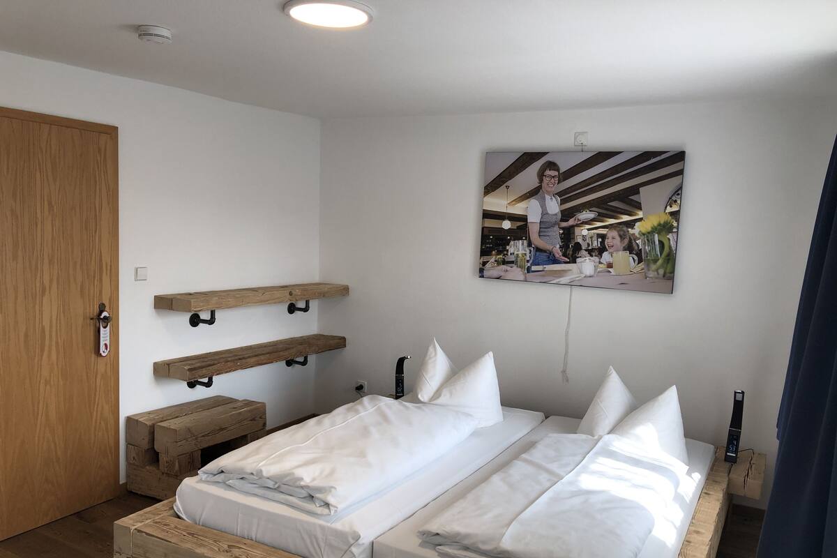 Een tweepersoonsbed met witte lakens, een houten hoofdbord en nachtkastjes. Er zijn houten planken aan de muur, een schilderij en een digitale klok.