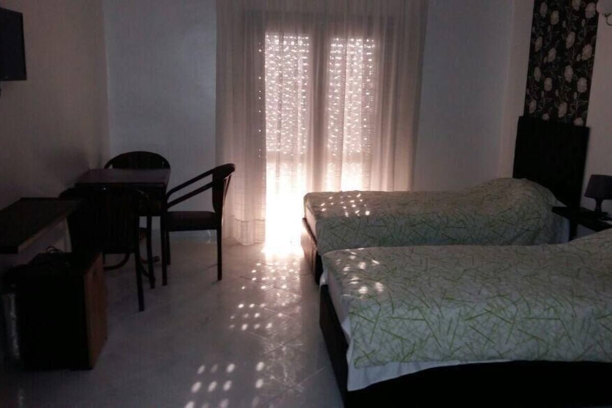 A hotel room with two single beds, a desk with a chair, a TV, and a window with sheer curtains.