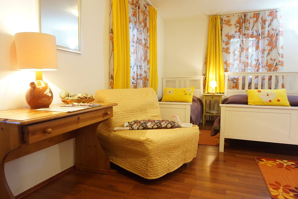 A cozy room with a wooden desk, a comfortable chair, a bed with yellow bedding, and floral curtains.
