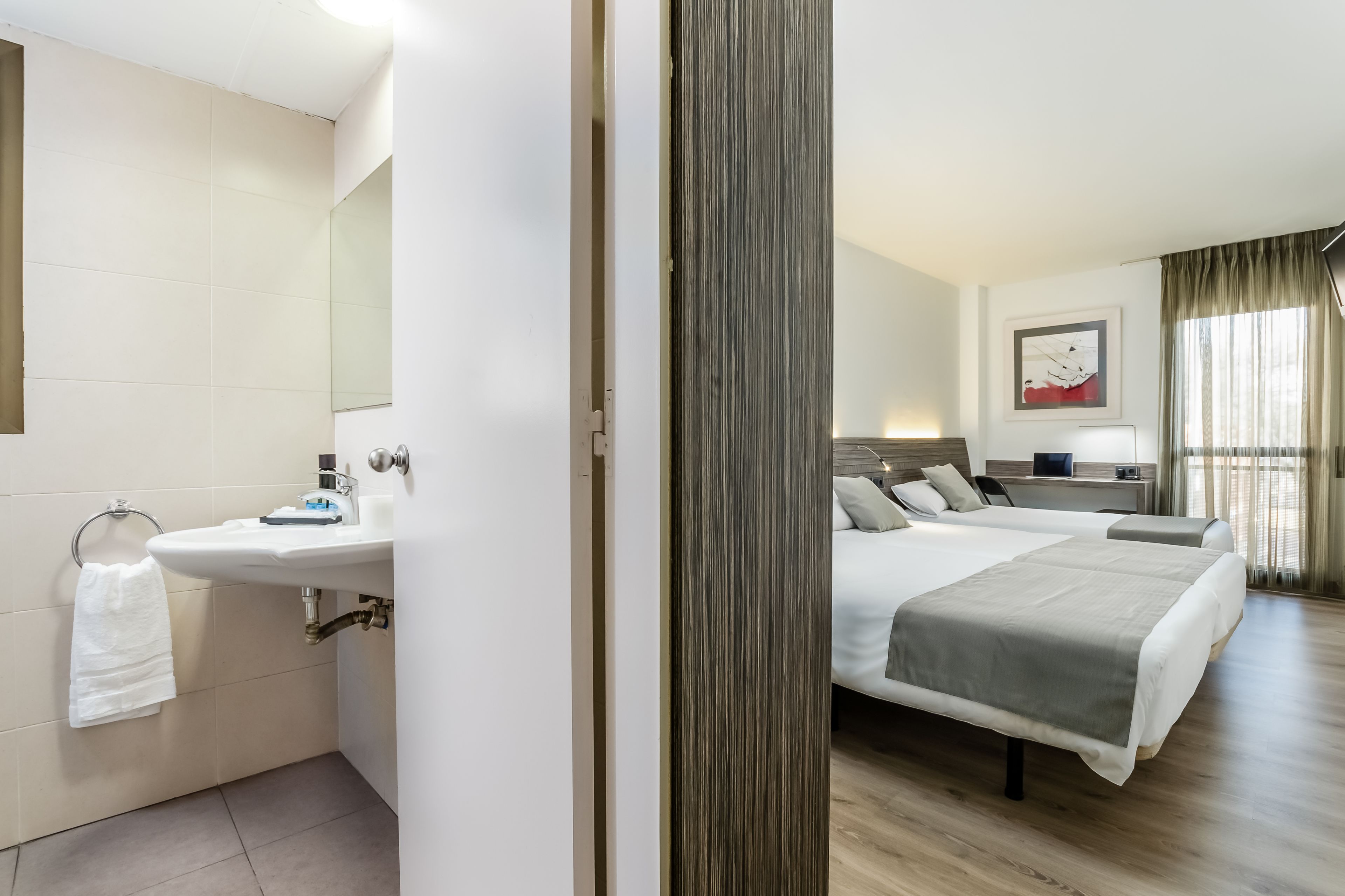 Habitación de hotel con cama, mesita de noche, lámpara y vistas al exterior a través de una ventana.