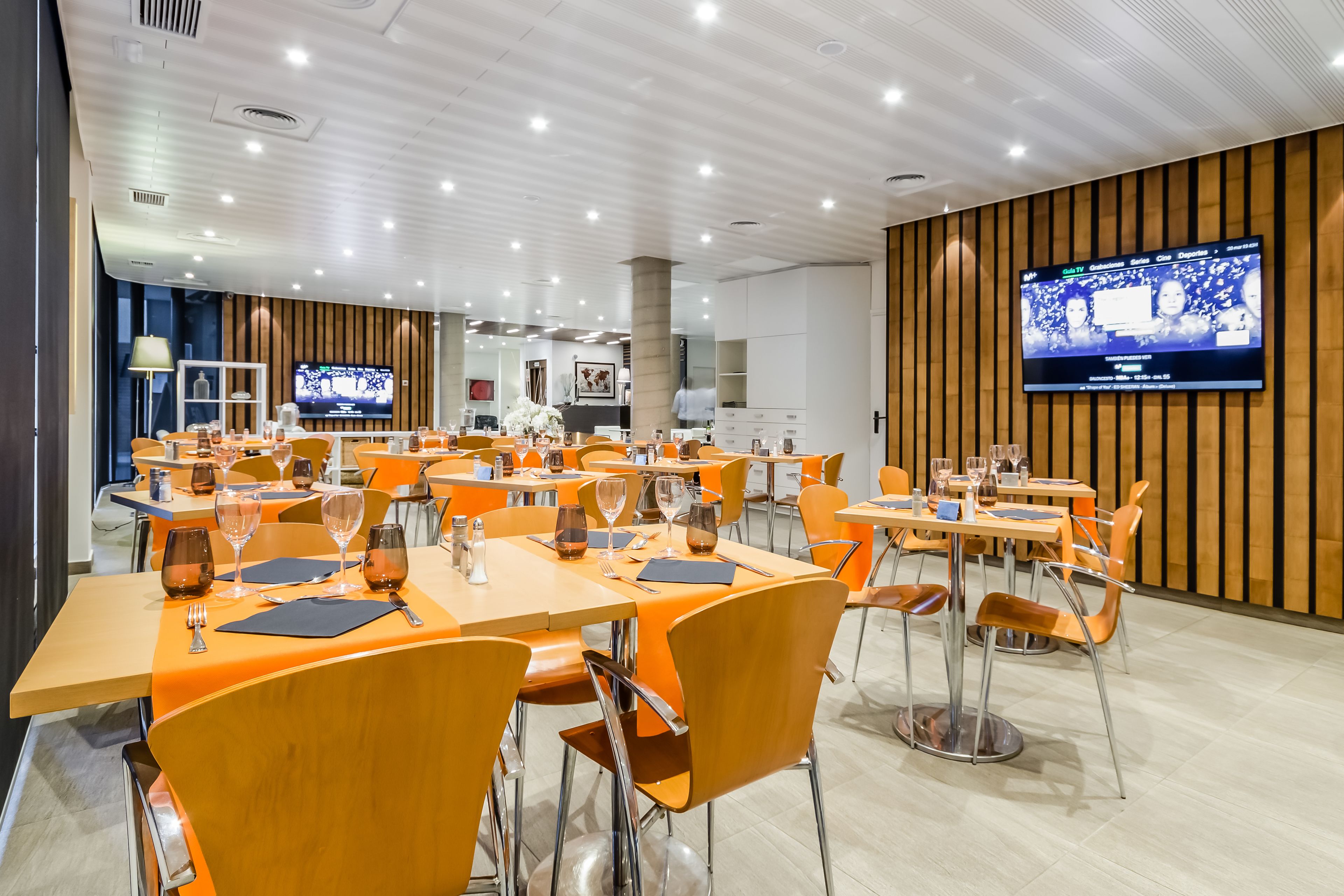 A modern dining area with multiple tables and chairs, a television screen on the wall, and a clean, well-lit interior.