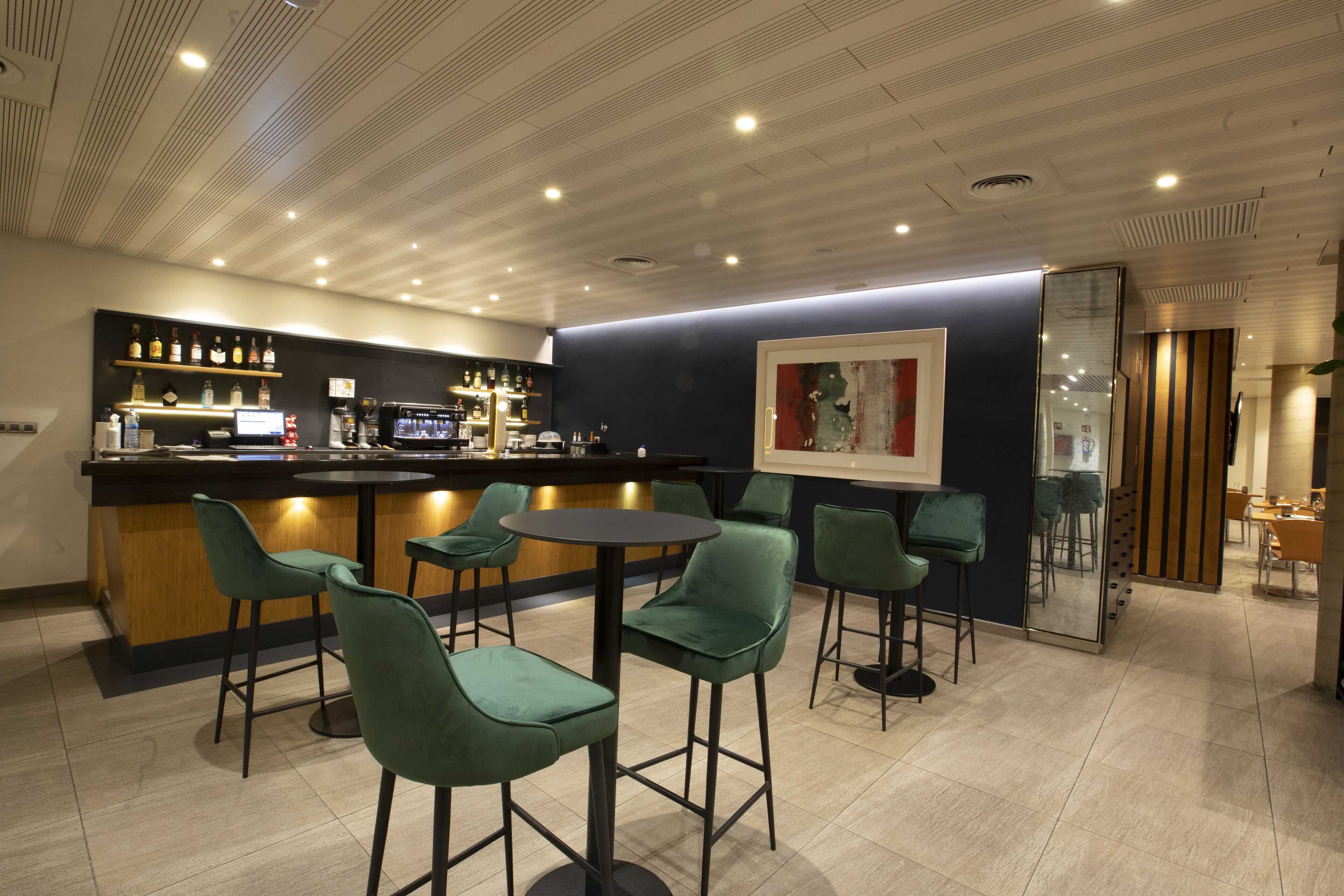 Zona de bar moderna con mostrador, taburetes y un área de estar con mesas redondas y sillas.