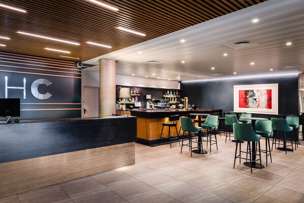 Un área de bar moderna con mostrador de recepción, taburetes y una barra con pantalla digital.