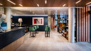 A modern interior with a reception desk, green bar stools, a large abstract painting, and a bookshelf filled with books and plants. - Hotel HC Mollet Barcelona (Mollet del Valles)