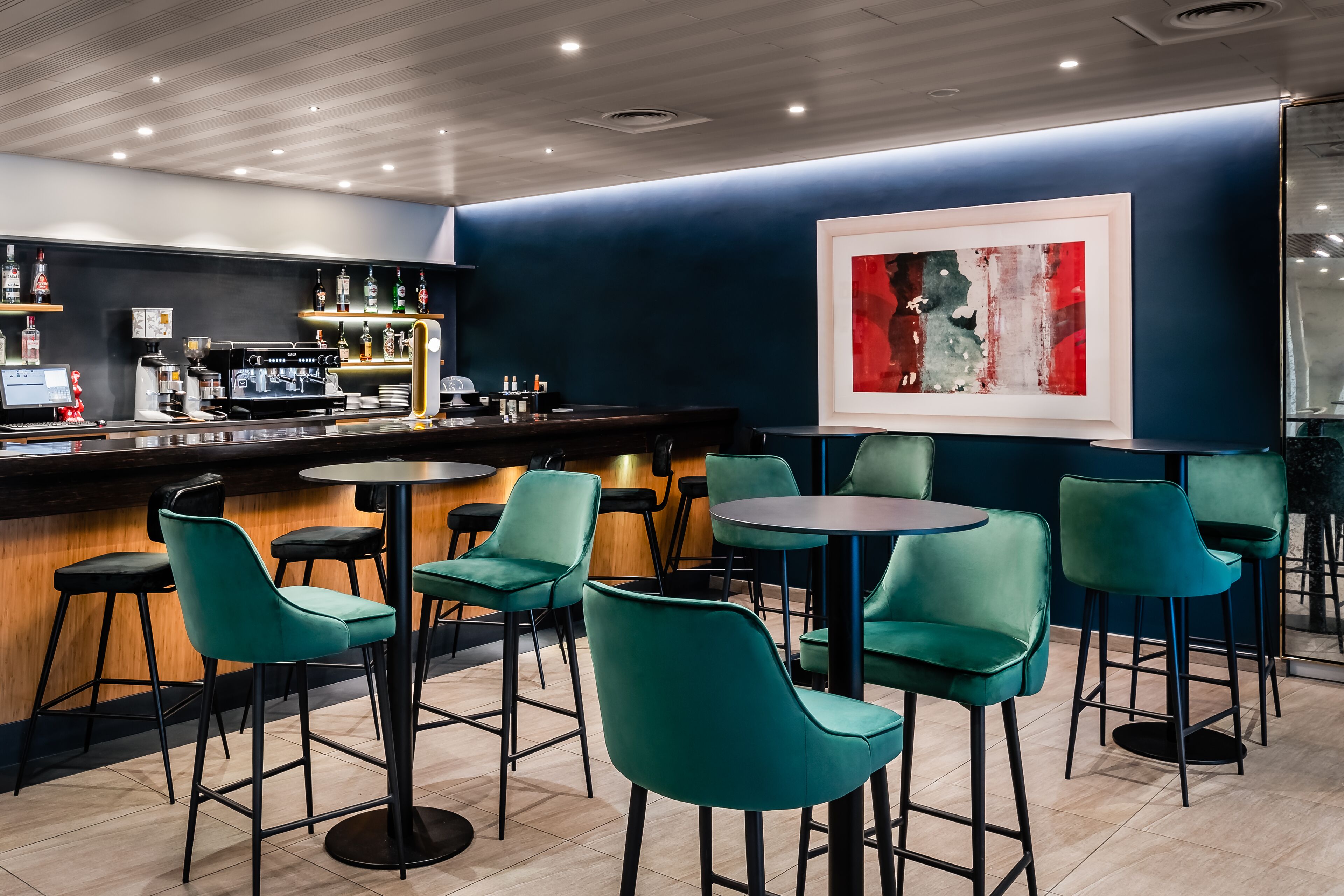 A bar area with teal chairs, a counter with a coffee machine, and a framed abstract painting on the wall.