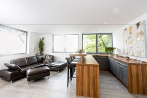 A modern kitchen with a wooden island, dark leather sofa, and a large window with a view of greenery. - Postsattler City Apartments (Albstadt)