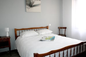 Un letto rifatto con lenzuola bianche, una testiera in legno e un solo cuscino. Una rivista è posata sul letto. Sul lato sinistro del letto, un comodino con una lampada.