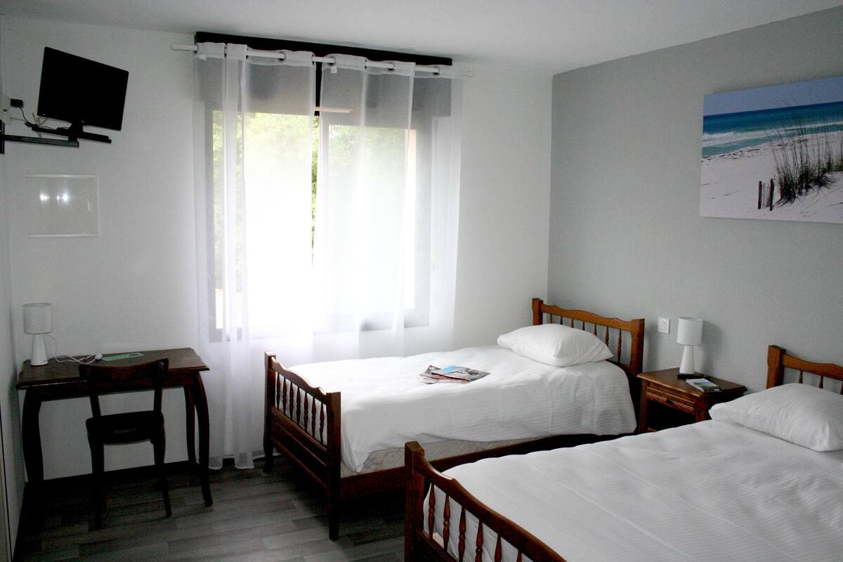 A hotel room with two single beds, a desk with a chair, a television mounted on the wall, and a window with sheer curtains.