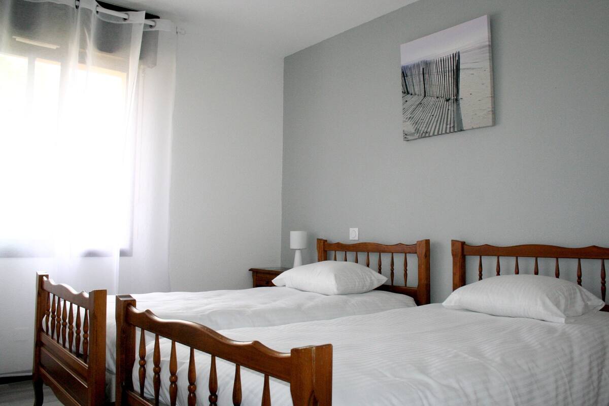 A room with two wooden beds, white bedding, a wall-mounted artwork, and a window with sheer curtains.