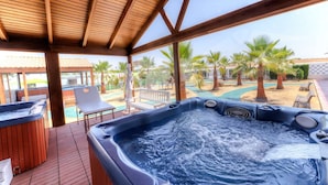 A hot tub area with a wooden deck, a pool, and palm trees in the background. - Cortijo Los Naranjos de San Juan (Las Cabezas de San Juan)