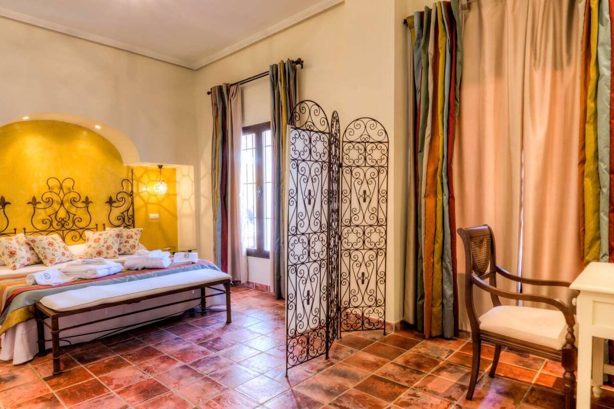 Une chambre à coucher comprenant un lit, une chaise, une table de chevet, un élément de séparation décoratif et une fenêtre avec des rideaux.