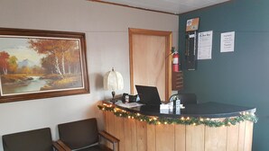 Un comptoir d’accueil avec un ordinateur portable, un extincteur et un tableau encadré. On aperçoit deux chaises et une porte à l’arrière-plan.