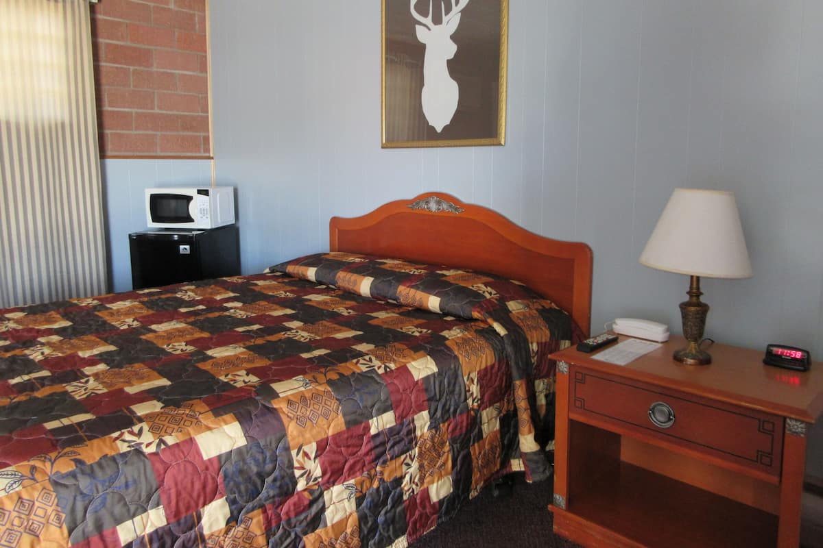 A hotel room with a bed, a nightstand, a lamp, a clock, a microwave, and a television.