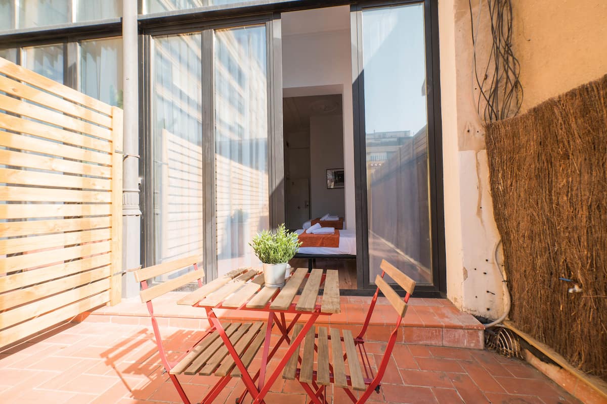 Una pequeña zona de estar al aire libre con una mesa y sillas de madera, una planta en maceta y una vista parcial de una habitación con cama y ventana.