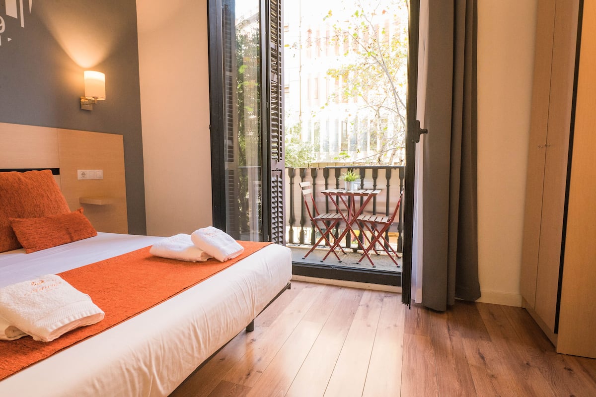 Habitación de hotel con cama, balcón con mesa y sillas, y vistas de la ciudad.