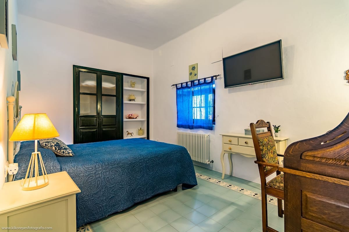 A bedroom with a bed, a desk, a chair, a TV, and a window with curtains.