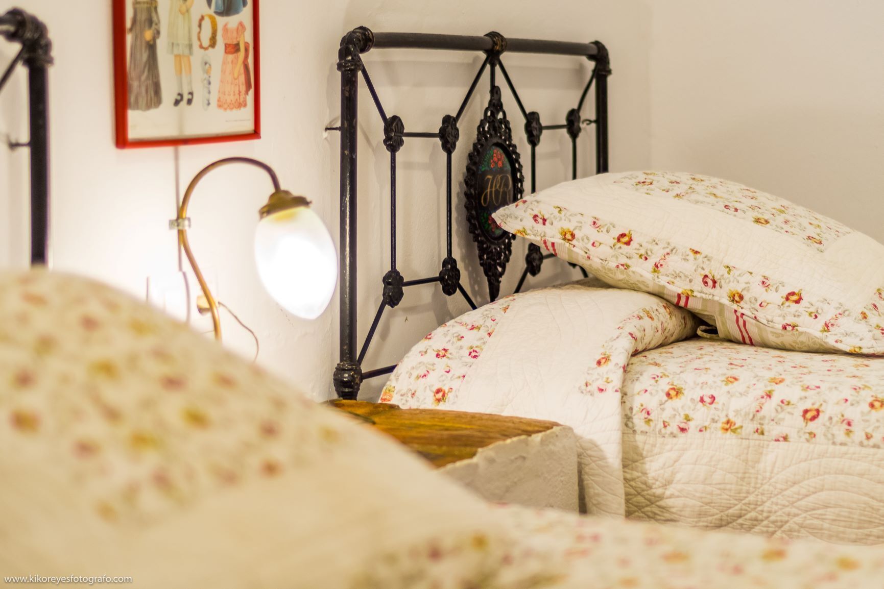 Un lit avec une literie à motifs floraux, une tête de lit en métal, une lampe fixée au mur et un tableau encadré.