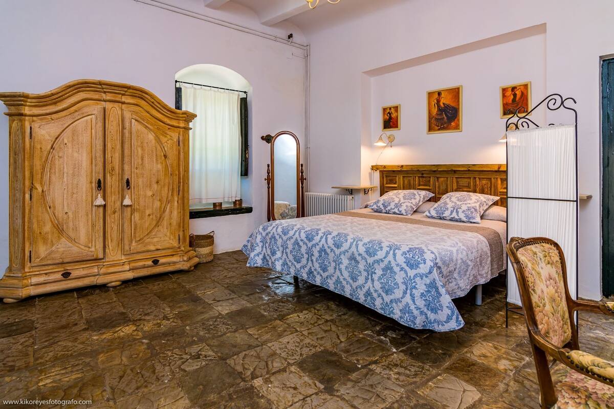 A bedroom with a large wooden wardrobe, a bed with patterned bedding, a radiator, and a chair.