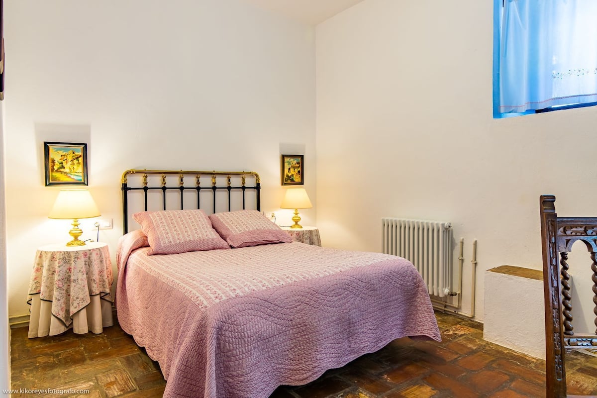 A bedroom with a bed, two bedside lamps, a radiator, and a wooden chair.