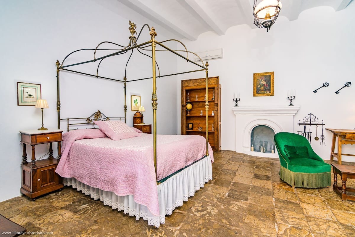 A four-poster bed with a white and pink bedspread, a wooden nightstand with a lamp, a green armchair, a fireplace, and a bookshelf with decorative items.