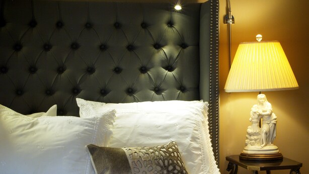 A neatly made bed with white linens and decorative pillows, a tufted headboard, a bedside lamp, and a small table with a statue. - Old Ivy Walk (Niagara-on-the-Lake)
