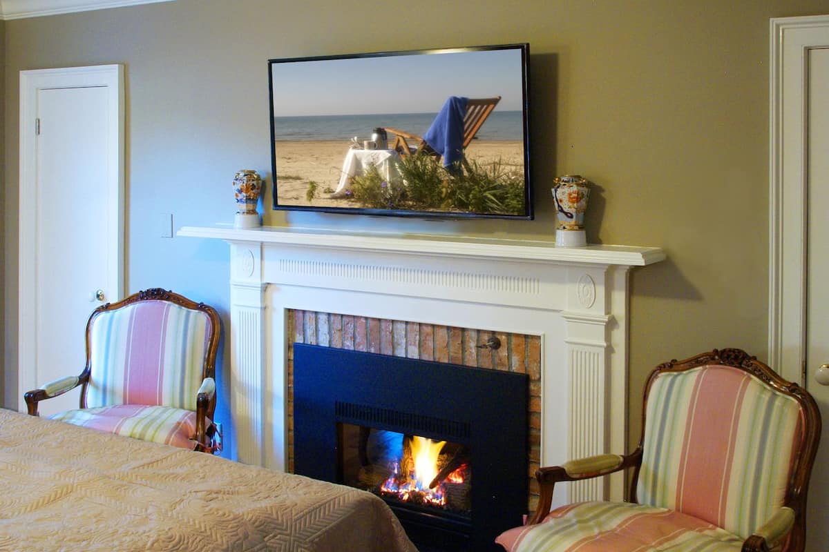 A room with a fireplace, two striped armchairs, a television mounted above the fireplace, and a bed with a patterned bedspread.