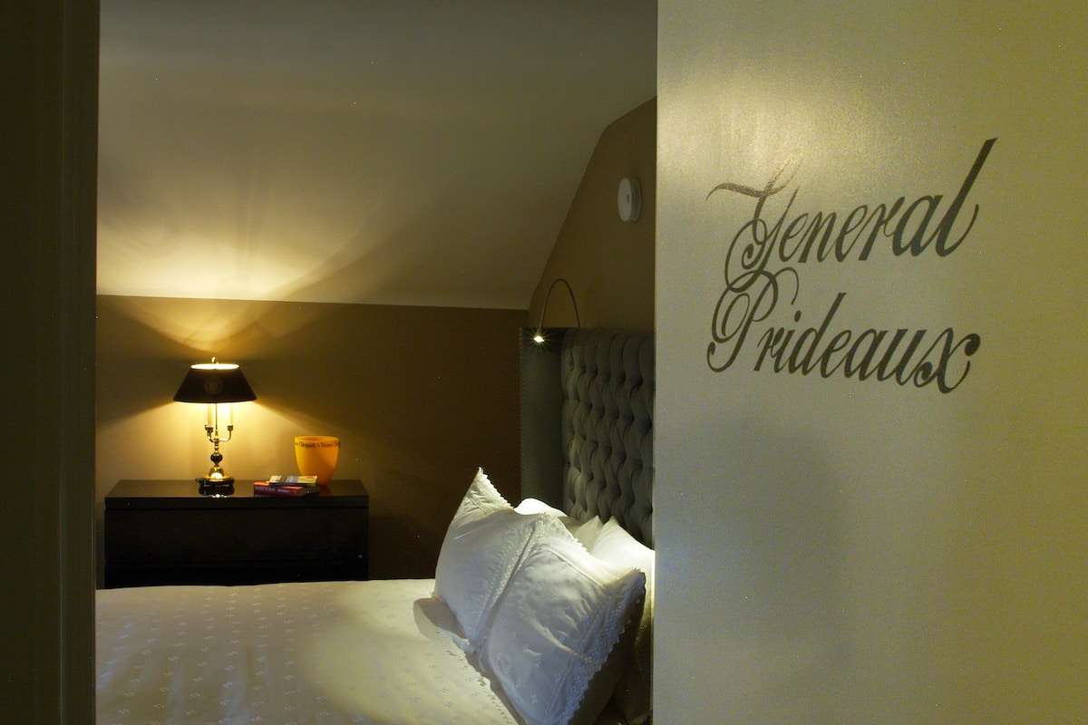A bedroom with a bed, a nightstand, a lamp, and a wall with the text "General Pradeaux".