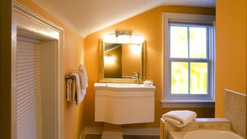 A bathroom with a white sink, mirror, and bathtub. There is a window with blinds and a towel rack with towels.
