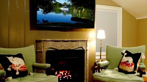 A living room with two green armchairs, a fireplace, a television mounted on the wall, and a window with blinds. - Old Ivy Walk (Niagara-on-the-Lake)