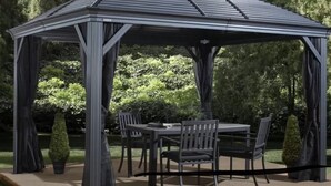 A gazebo with a metal frame and a roof, surrounded by chairs and a table, set in a garden with trees and shrubs. - Old Ivy Walk (Niagara-on-the-Lake)