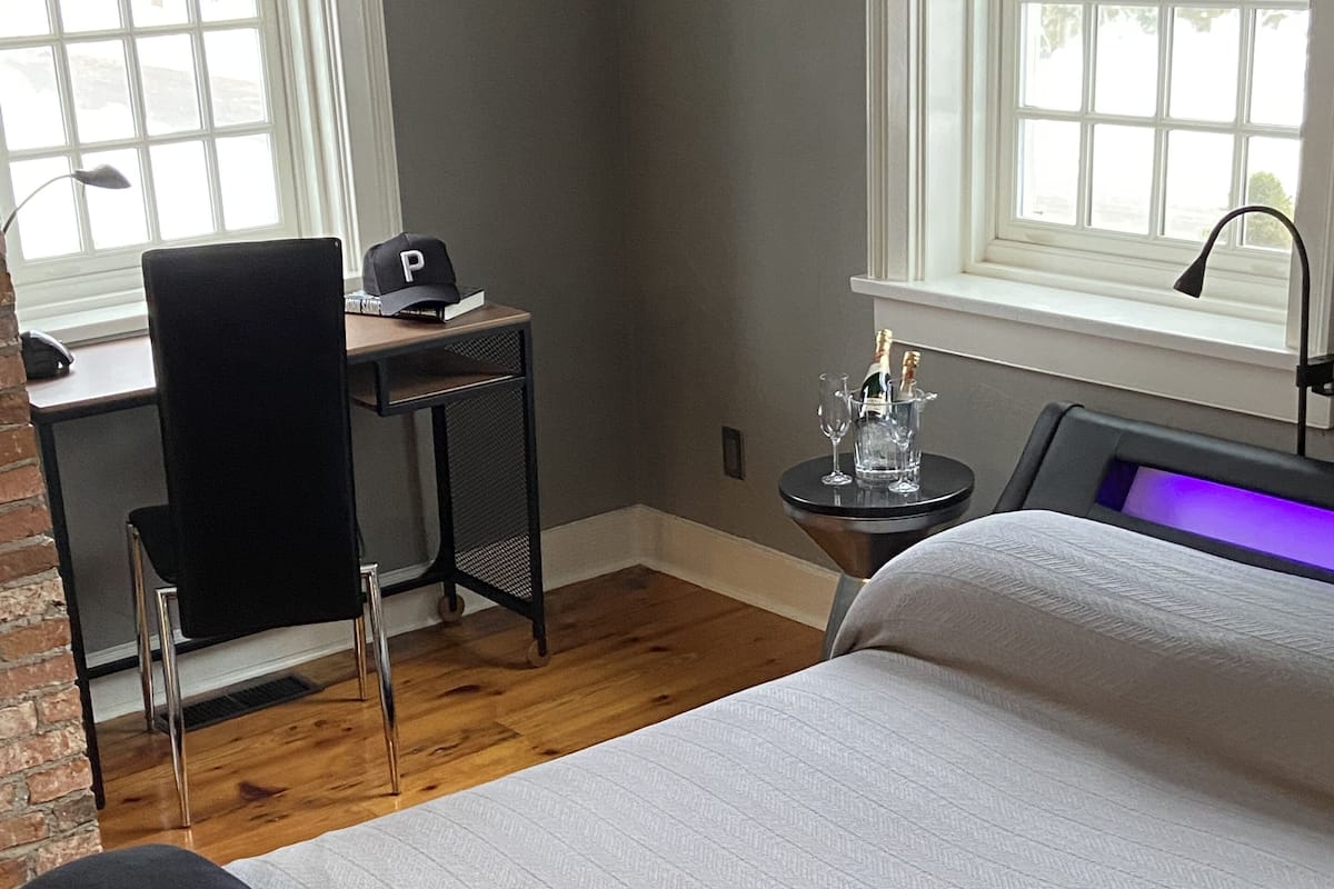 A bedroom with a bed, a desk, a chair, a small table with a bottle and glasses, and a window with a view of trees.