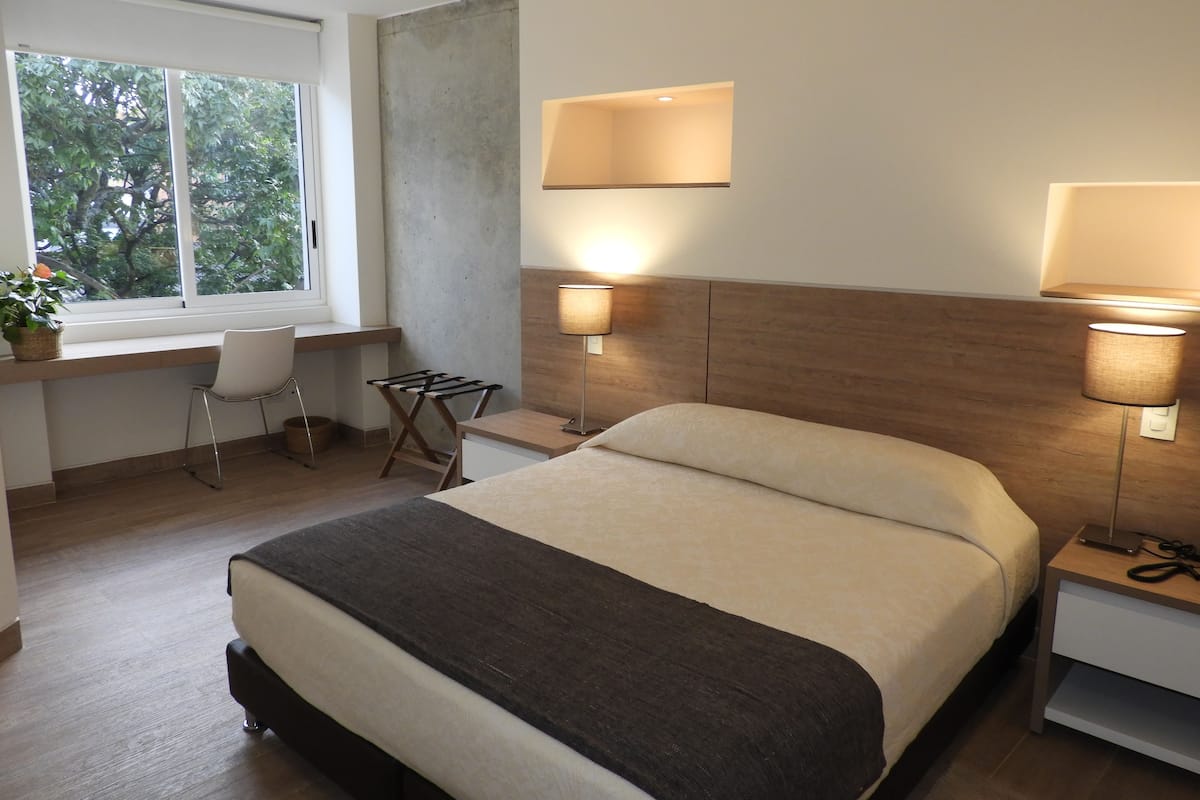 A modern hotel room with a large bed, a desk with a chair, two wall-mounted lamps, and a window with a view of trees.