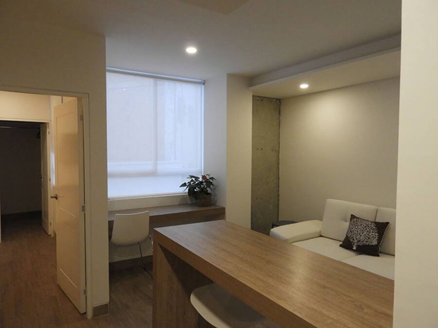 Ein modernes, minimalistisches Zimmer mit einem Holztisch, einem weißen Sofa mit Zierkissen, einer Topfpflanze und einem großen Fenster, das natürliches Licht hereinlässt.