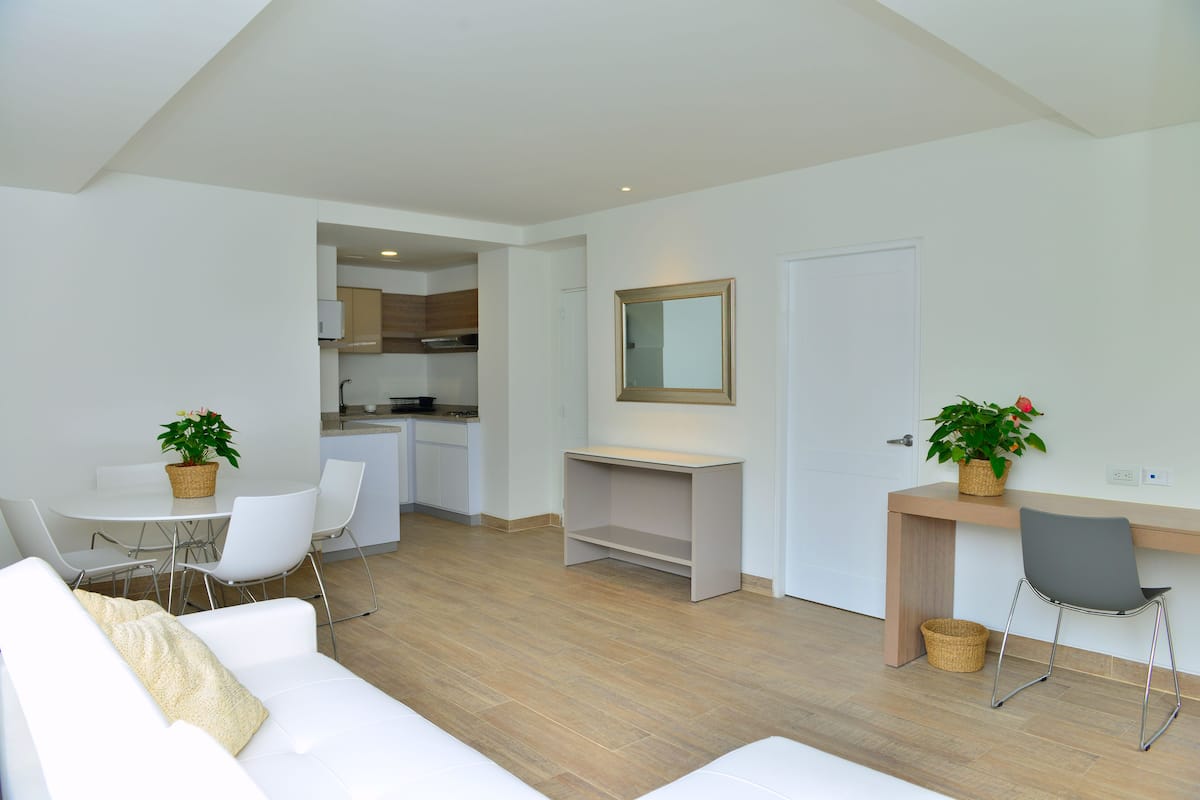 A modern living room with a white sofa, a dining table with chairs, a kitchen area, and a small desk with a chair.