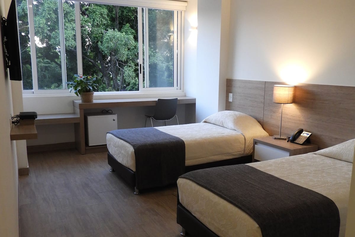 A hotel room with two beds, a desk, a chair, a lamp, and a window with a view of trees.