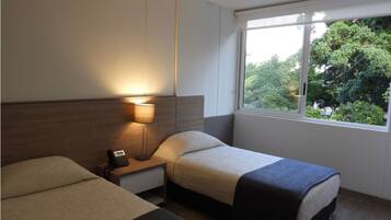 A hotel room with two beds, a nightstand with a lamp, a window with a view of trees, and a small rug on the floor.