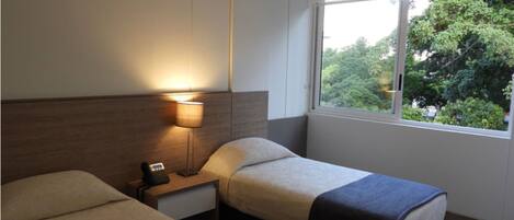 A hotel room with two beds, a nightstand with a lamp, a window with a view of trees, and a small rug on the floor.