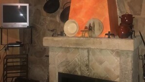 Een stenen openhaard met hangende potten, een televisie op een standaard en een tafel met een kleurrijke tafelkleed.