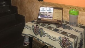 Een kamer met een trap, een lijst met een afbeelding aan de muur, een tafel met een bloemrijk tafelkleed en een waterfles.