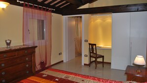 A bedroom with a wooden dresser, a bed with a red and beige bedspread, a chair, and a nightstand with a lamp. - Tenuta Fortelongo (Fara Vicentino)