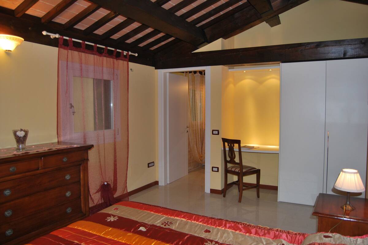 A bedroom with a wooden dresser, a bed with a red and beige bedspread, a chair, and a nightstand with a lamp.