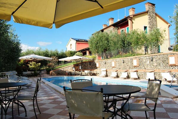 A poolside area with outdoor seating and umbrellas, a stone wall, and a building with a red roof in the background. - Tenuta Fortelongo (Fara Vicentino)