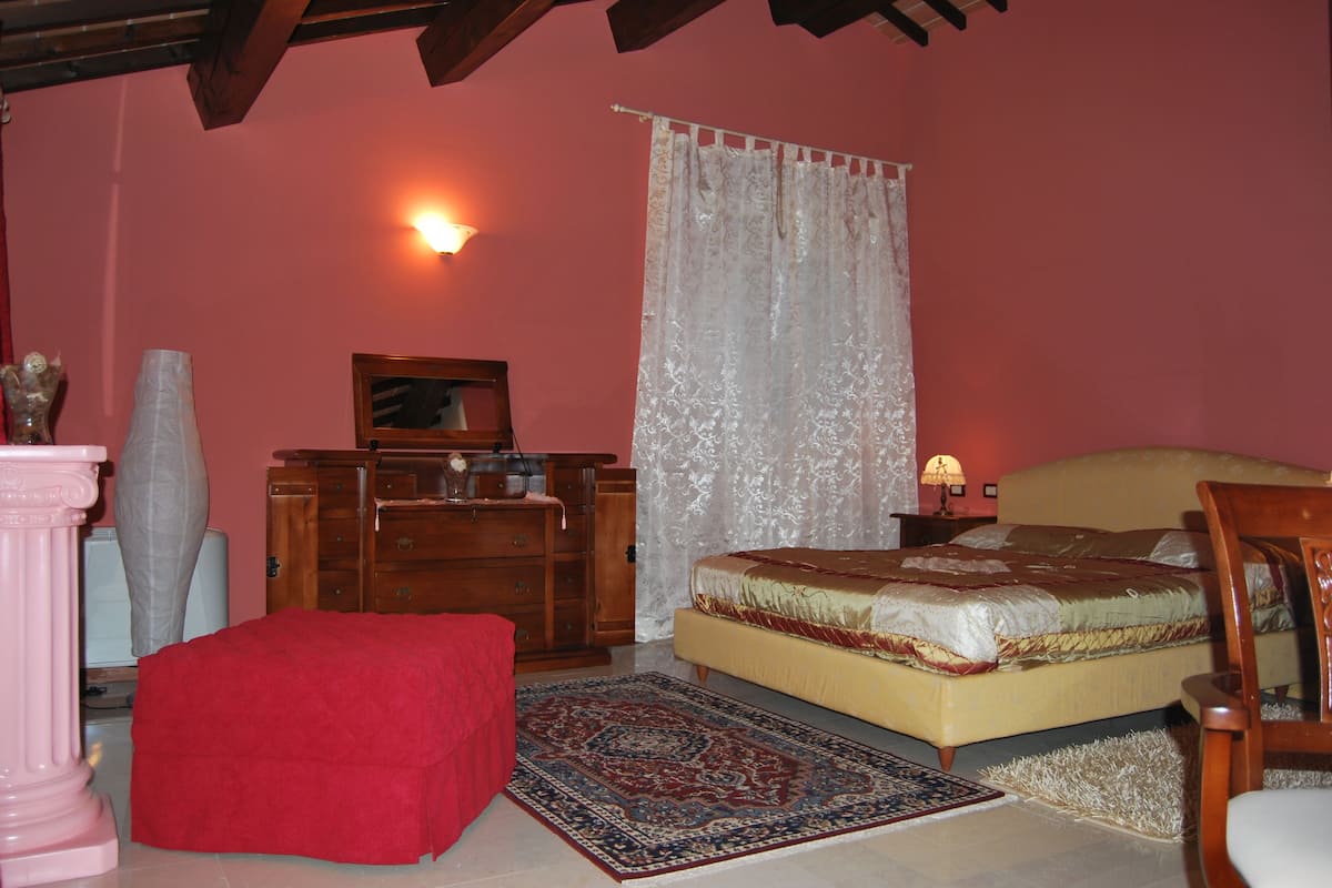 A bedroom with a bed, a wooden dresser, a red ottoman, a nightstand with a lamp, and a large window with a white curtain.