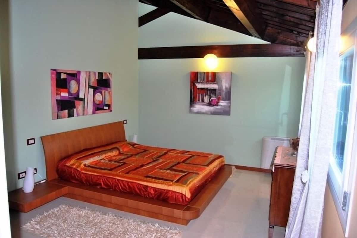 A bedroom with a bed, two paintings, a nightstand, and a window.