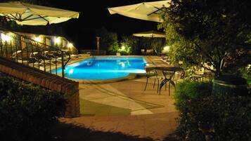 A nighttime outdoor swimming pool area with lounge chairs and umbrellas, surrounded by greenery.
