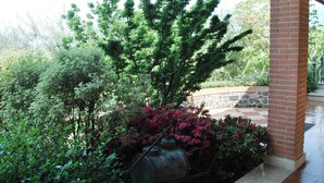 A covered outdoor area with a brick column, a metal barrel, and various green shrubs and pink flowers. - Tenuta Fortelongo (Fara Vicentino)