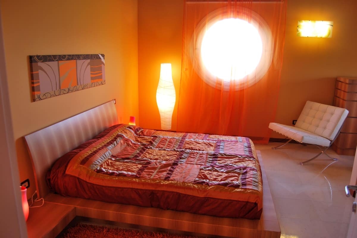 A bedroom with a bed, a chair, a wall-mounted lamp, and a window with curtains.