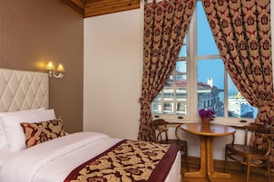 A hotel room with a bed, two bedside lamps, a window with curtains, a small table with a chair, and a view of buildings and a boat outside.